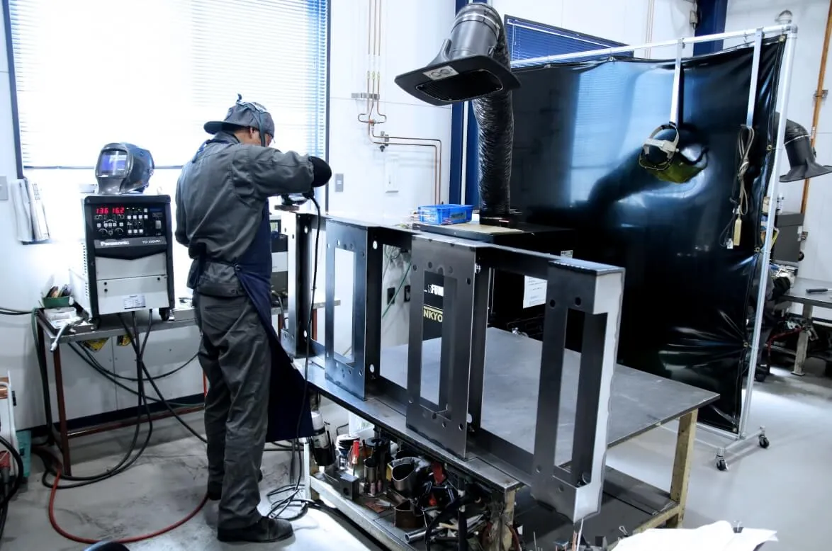 作業着を着た技術者が、金属フレームを研磨機で加工している様子。